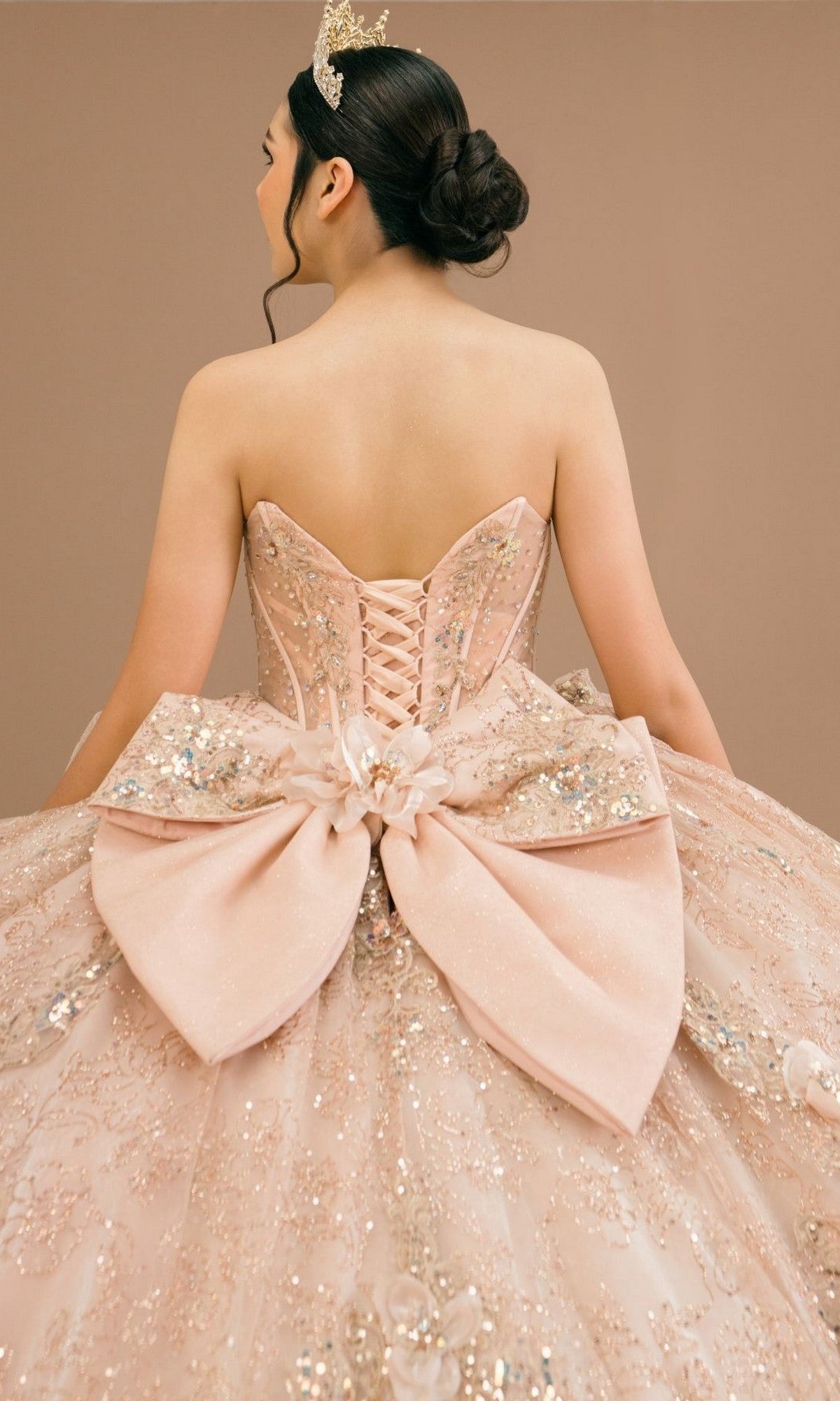 A woman is seen from behind against a neutral background, wearing the Quinceanera Dress 8590JQ By Cinderella Couture—a pink, glittery ball gown featuring a beaded corset bodice and a large bow at the back. She completes the look with a tiara.